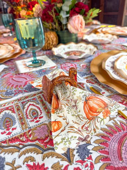A Turkey & Gourds Guest Towels/20pk harvest pattern napkin decorated with pumpkins and autumn leaves, tucked into a turkey-shaped napkin ring on a patterned tablecloth.